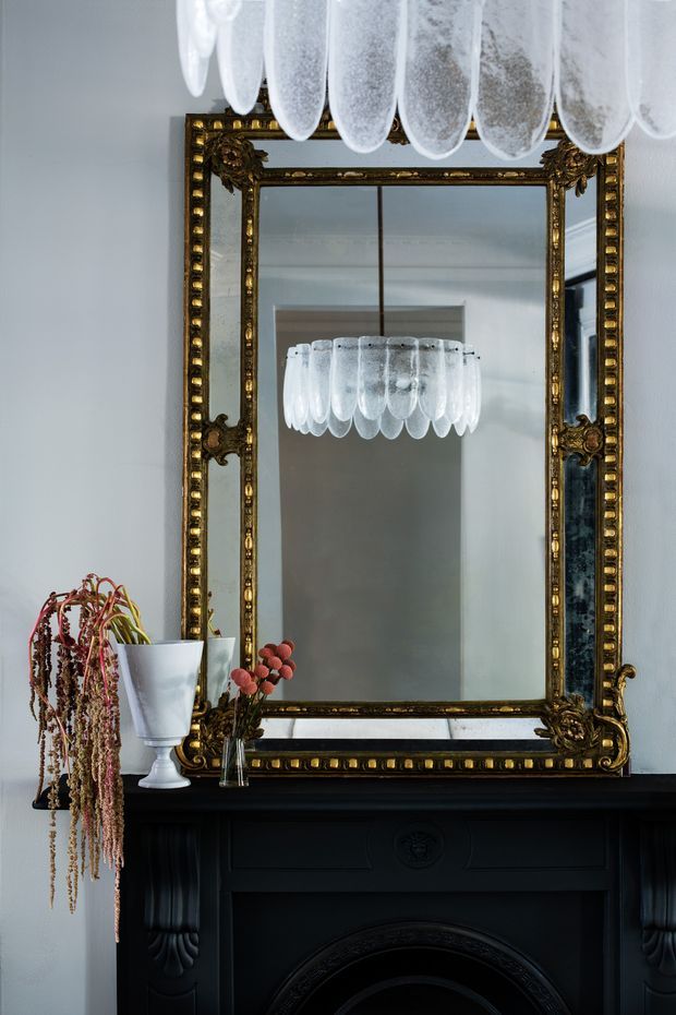 A refined interior vignette centred on an ornate antique gold-framed wall mirror above a dark, elaborately detailed fireplace mantel. A sculptural crystal pendant light is reflected in the mirror, while a white vase and dried floral arrangement add decorative detail. The room has pale grey walls, strong black architectural joinery, and a formal, heritage-inspired atmosphere.