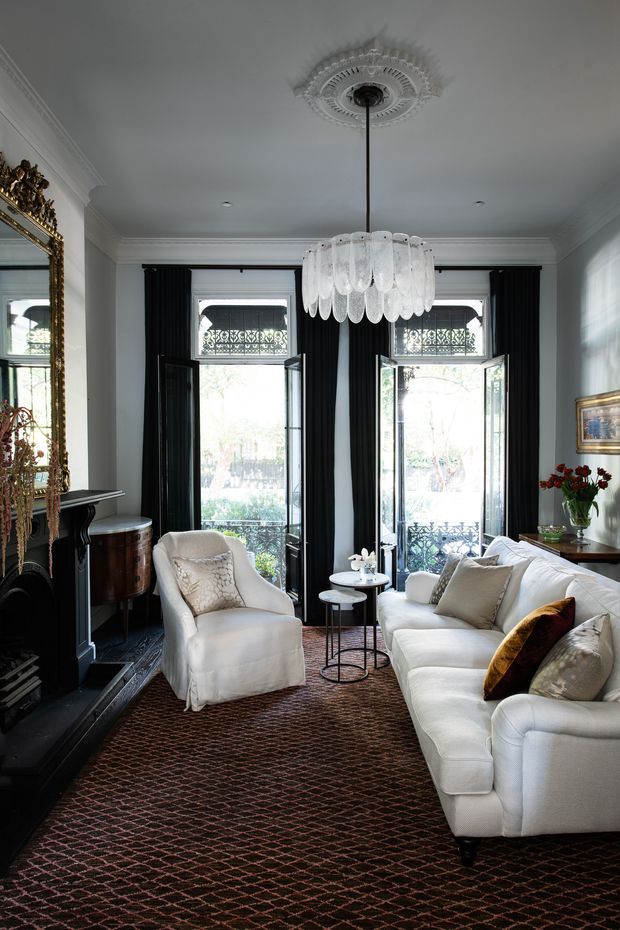 Elegant traditional living room with white upholstered seating, a black fireplace surround, ornate gold-framed mirror, dark curtains, tall French doors, decorative ceiling medallion and crystal chandelier. The room has a refined, layered palette of white, black, dark brown and gold, with patterned carpeting and classic mouldings.