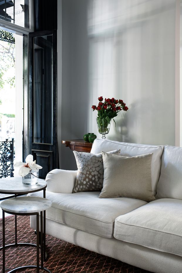 A refined living room corner with a large cream upholstered sectional sofa, layered neutral cushions, nesting round coffee tables, and a dark patterned rug. A dark timber-framed glazed door opens to a balcony, while a timber console with a vase of red flowers sits against a tall pale wall. Strong natural light creates soft shadows across the wall, giving the room an elegant, calm atmosphere.