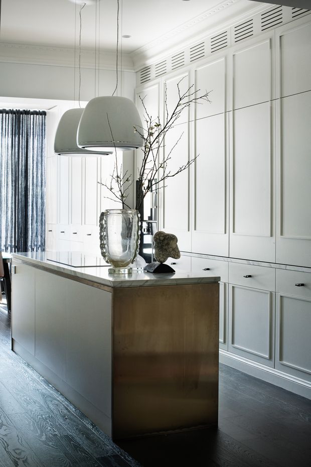 A refined, monochromatic kitchen with a large central island, white panelled cabinetry, integrated louvred ventilation panels, dark timber flooring, and a pale stone benchtop. Oversized sculptural pendant lights hang above the island, while a bronze-toned island end panel and decorative vase provide subtle contrast.