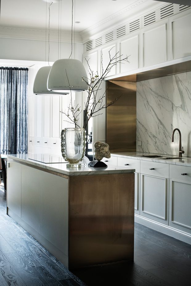 A refined, light-filled kitchen with a large central island, white raised-panel cabinetry, marble splashback and benchtops, and a brushed metal island end panel. Architectural details include decorative wall panelling, integrated louvred cabinet ventilation panels, pendant lights, and dark timber flooring. The palette is predominantly white and grey with warm metallic accents.