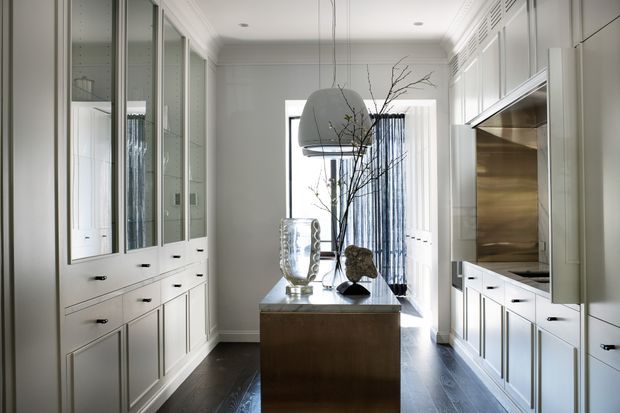 A refined, narrow kitchen with white built-in cabinetry and glass-front display cupboards lining both sides. A central dark timber island or console anchors the room beneath a large pendant light, while stainless steel appliances and a reflective metallic splashback occupy the right-hand wall. Dark timber flooring, pale walls, decorative moulding, and a tall window with sheer curtains create a bright, elegant interior.
