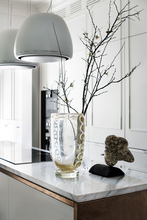 A refined, bright kitchen interior featuring a white marble island with waterfall-style edging, white panelled cabinetry, black integrated appliances, and large sculptural white pendant lights. A clear glass vase with bare flowering branches and small stone-like decorative objects provide understated styling. The space has a polished, monochromatic contemporary look with classic panel detailing.
