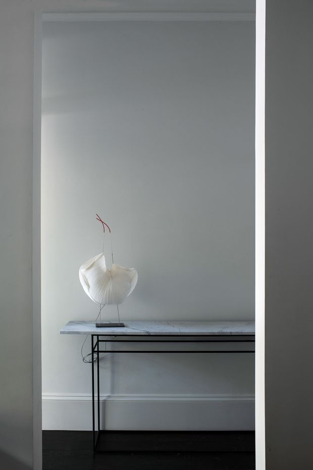 A minimalist interior vignette viewed through a doorway, featuring a slim black metal console table with a pale stone or marble-look top against a light grey wall. A sculptural white pendant or decorative light sits on the console, creating a quiet, refined composition. Dark timber flooring and simple white wall trim provide contrast.