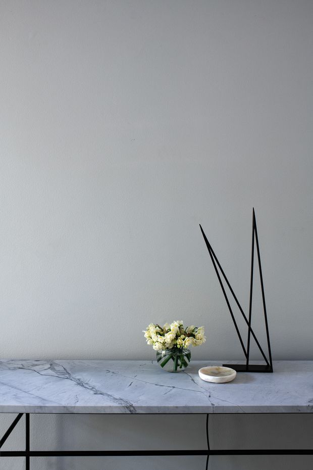 A minimalist interior vignette featuring a slim black metal console table with a veined white marble top, set against a light grey textured wall. A small glass vase of white flowers and a shallow white decorative dish sit on the console, alongside a tall angular black metal sculptural object. The composition is sparse, calm, and refined.