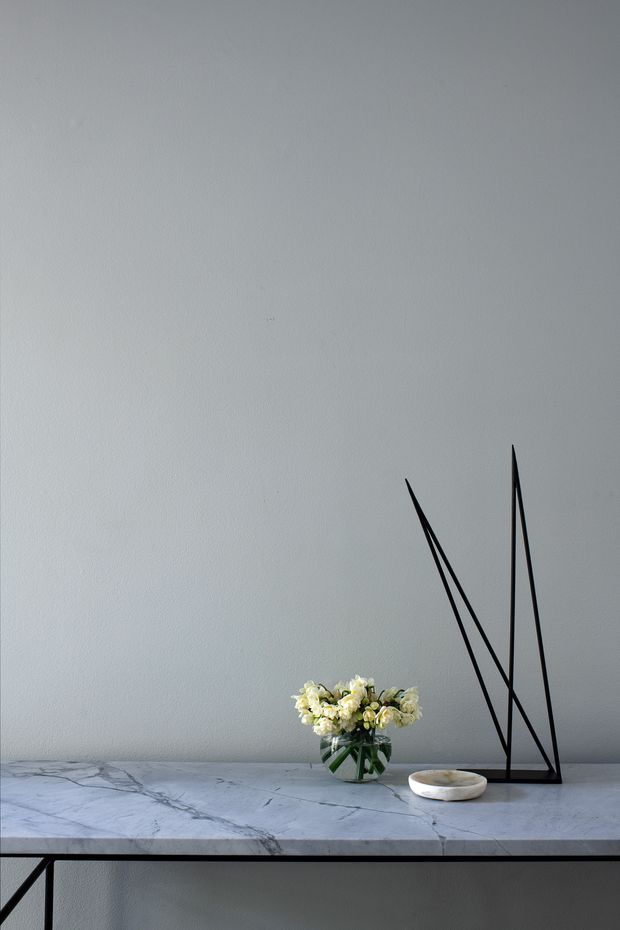 A minimalist interior vignette featuring a pale grey textured wall, a slim black console table with a white-grey veined marble top, a small glass vase of white flowers, and a simple black geometric sculpture. The composition is subdued, refined, and heavily focused on negative space and material contrast.