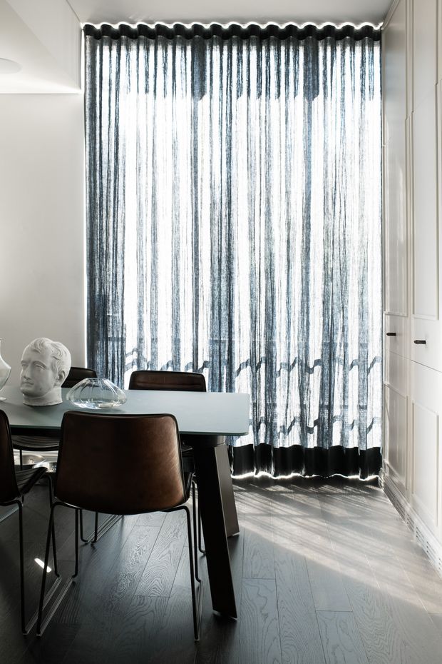 A refined dining area with a dark rectangular dining table and brown leather chairs, positioned in front of full-height sheer charcoal curtains. White paneled cabinetry or wall joinery lines the right side, while pale timber flooring and strong filtered daylight create a high-contrast, elegant interior. Decorative glassware and a sculptural bust sit on the table.