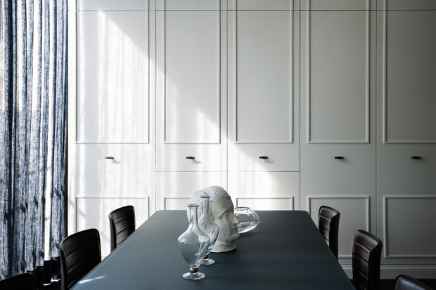 A refined dining room featuring a long dark dining table with sculptural glassware and pale decorative vessels, surrounded by dark upholstered dining chairs. The room has white decorative wall panelling with framed recessed sections and small dark handles, alongside full-height sheer textured curtains. Strong natural light creates a sophisticated, high-contrast atmosphere.