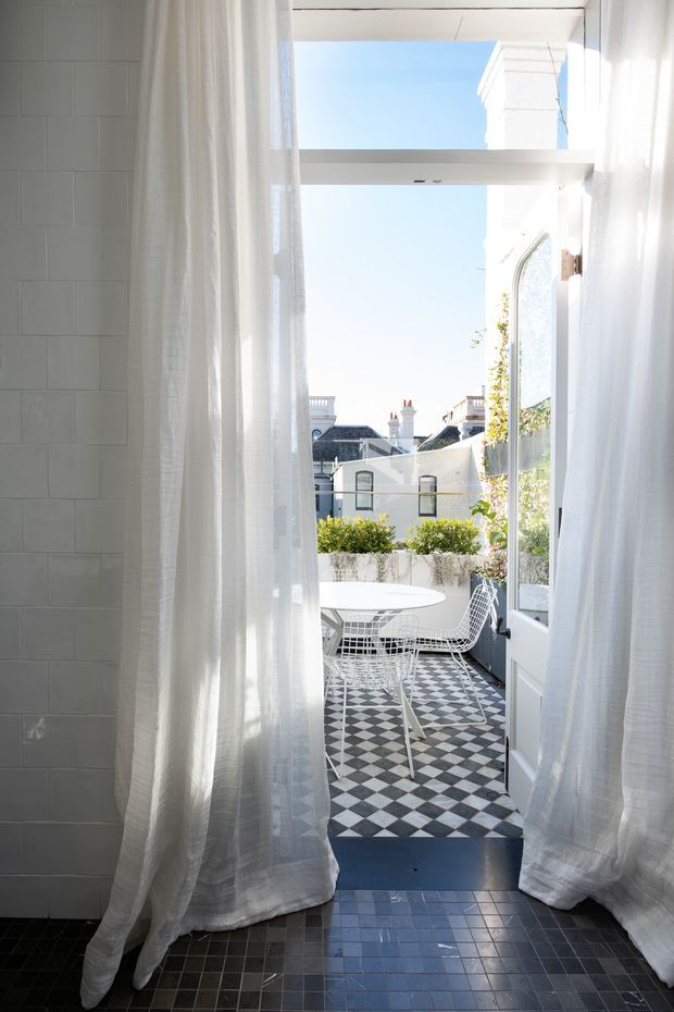 Bright view from an interior tiled space through open French doors onto a small rooftop terrace or balcony. Sheer white curtains frame the opening, while the terrace has a bold black-and-white checkerboard tile floor, white wire-frame dining chairs, a round white table, and raised planter greenery. White rendered neighbouring buildings and a clear blue sky are visible beyond.