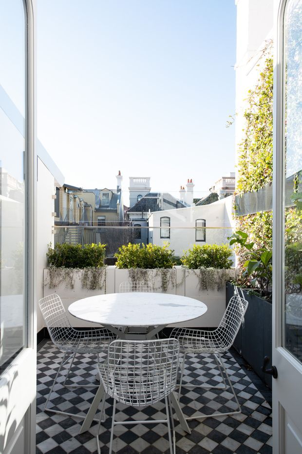 Bright, narrow outdoor terrace or balcony accessed through open glazed doors, furnished with a round white dining table and wire-frame chairs. The terrace has a striking black-and-white checkerboard tiled floor, raised planters with greenery, and views toward closely spaced ornate urban residential buildings. The space feels airy, refined, and classically urban.