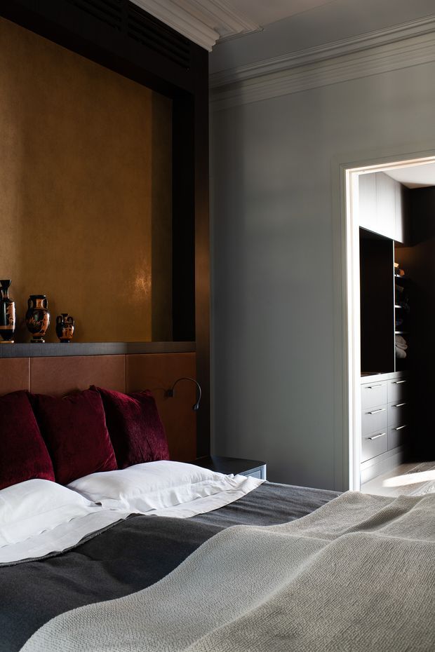 A moody, refined bedroom with a low upholstered headboard, deep burgundy pillows, layered grey and white bedding, and a warm metallic ochre feature wall framed in dark trim. Decorative ceiling cornices and wall mouldings add classical detailing. An open doorway reveals dark built-in cabinetry and drawers in an adjoining dressing or storage area.
