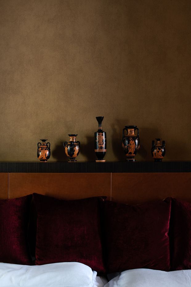A close-up, moody bedroom vignette featuring a dark upholstered or timber headboard, deep burgundy velvet pillows, crisp white bedding, and a black shelf displaying a collection of decorative ceramic vessels against a warm textured brown wall.