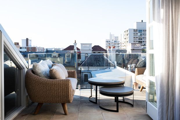 A bright urban apartment terrace or balcony furnished with a woven lounge chair, nested round coffee tables, and a second light-coloured chair beside full-height glazed doors. Pale tiled flooring and a glass balustrade frame expansive city and rooftop views beyond.