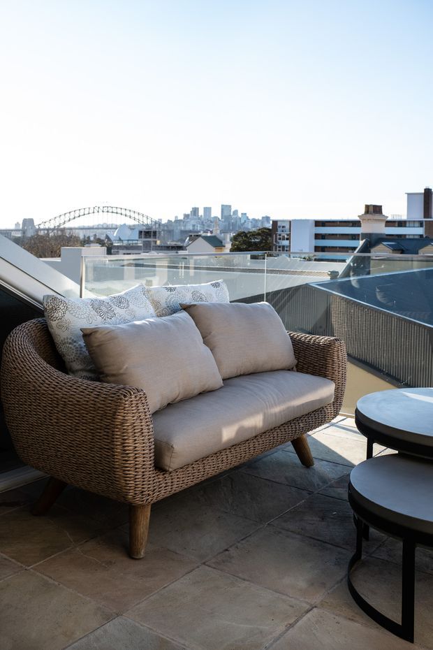 A bright rooftop terrace with a woven outdoor lounge chair, neutral cushions, and round black tables on large stone tiles. Clear glass balustrades frame an urban skyline with a prominent arched bridge in the background. The space is styled as a relaxed outdoor seating area with open city views.