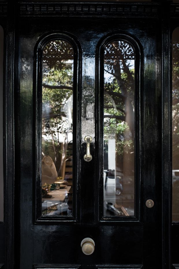 Close-up view of a glossy black, richly moulded entrance door with two tall arched glass panels, ornate decorative detailing at the upper glazing, and polished metal hardware. Reflections and views through the glass reveal a leafy courtyard or garden and a furnished interior beyond. The image emphasizes the door's heritage character, dark finish, and intricate craftsmanship.