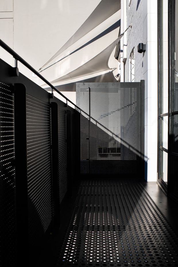 A narrow, contemporary outdoor walkway or balcony enclosed by black perforated metal privacy screens and a matching perforated metal floor. White masonry walls and tall glazed openings frame the space, while stretched triangular shade sails provide overhead cover. Strong sunlight creates patterned shadows through the perforations, producing a high-contrast, industrial architectural composition.
