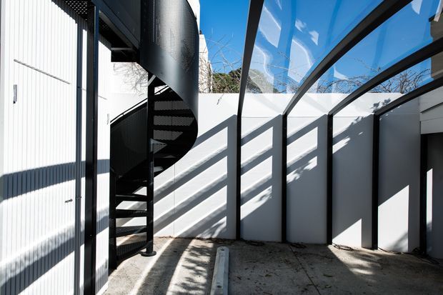 A bright contemporary exterior passage or courtyard beside a white corrugated-clad building. A black metal spiral staircase rises along the left side, while a black-framed transparent roof or covered walkway creates strong geometric shadows across the white walls and concrete floor.