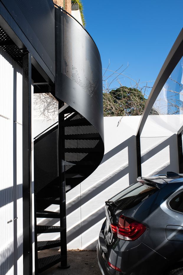 Exterior architectural view of a black perforated-metal spiral staircase attached to a contemporary building, with strong geometric shadows cast across a white rendered wall. A covered parking area and a parked car occupy the foreground, while blue sky and surrounding vegetation are visible above the courtyard walls.
