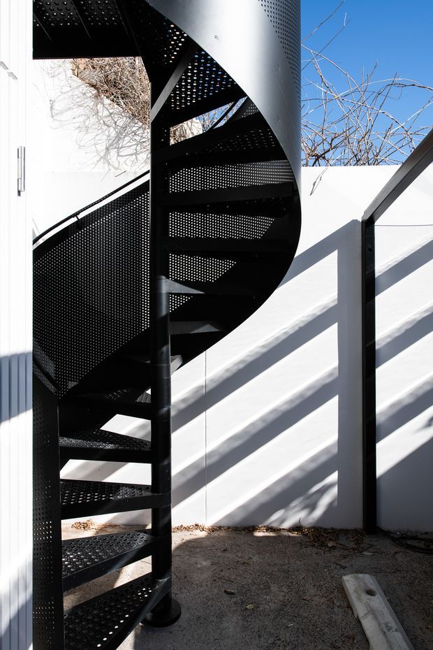 A compact exterior courtyard or access area featuring a striking black perforated metal spiral staircase. The staircase has open treads, a curved perforated guard and central support column, set against white rendered walls with strong angular sunlight and shadows. Bare branches are visible above the walls, and the ground is paved or concrete.