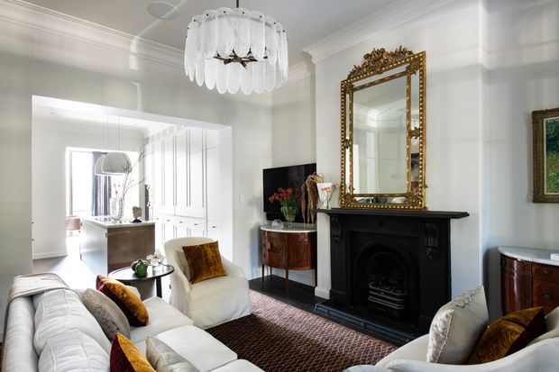 Elegant, bright formal living room with white upholstered seating, richly patterned cushions, a dark patterned rug, a black decorative fireplace, and an ornate gilt mirror above the mantel. The room features detailed ceiling cornices, white wall panelling, a sculptural pendant light, and an open connection to a bright kitchen or dining area.