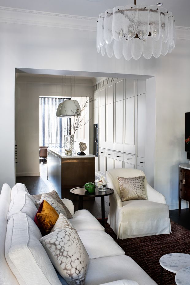 Bright, refined living room with a large white slipcovered sofa and armchair, patterned cushions, dark round coffee tables, and a dramatic layered glass pendant light. A wide opening leads to a kitchen or dining area with a dark island and extensive white built-in cabinetry featuring decorative panel moulding. The room has pale walls with traditional cornice detailing, dark timber flooring, and a patterned dark rug.
