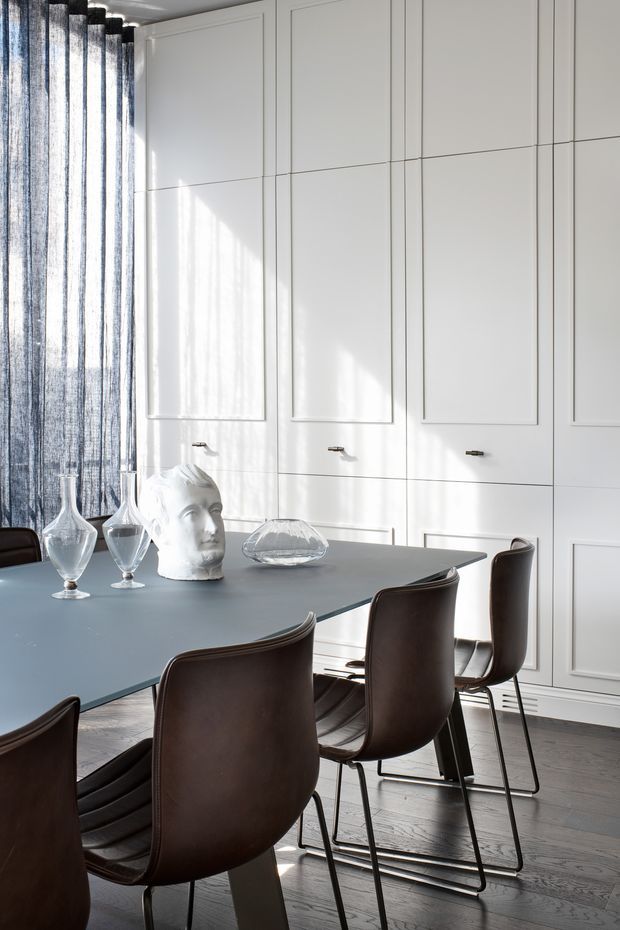A refined dining room with a large dark dining table surrounded by brown leather chairs. The room features full-height white paneled built-in cabinetry with discreet handles, dark timber flooring, sheer dark curtains, and sculptural glass and ceramic table decor. Strong sunlight creates a bright, elegant atmosphere.