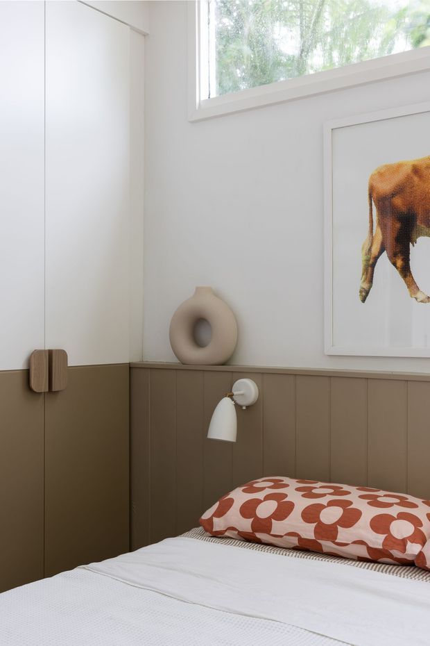 A compact, softly styled bedroom corner with white upper walls, taupe lower vertical wall panelling, built-in cabinetry, and a small wall-mounted bedside light. A bed with white bedding and a rust-red patterned cushion sits beneath framed artwork, with a sculptural ceramic vase on the ledge and daylight entering through a high window.