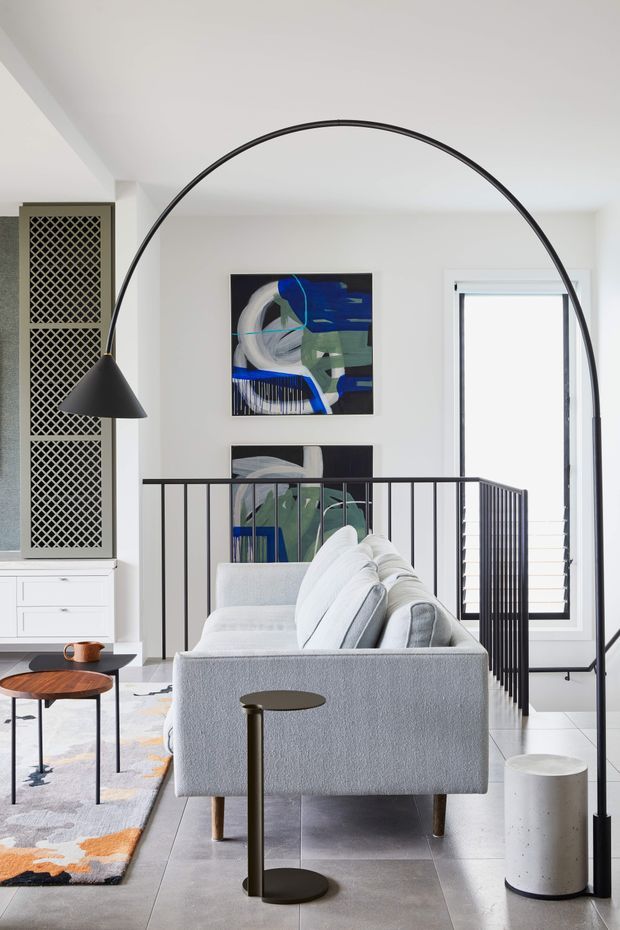 Bright contemporary living room with a light grey sofa positioned beside a black metal stair balustrade and dramatic arched floor lamp. The room features white walls and ceiling, large grey floor tiles, black-framed windows with blinds, abstract blue and green artwork, a patterned radiator screen or wall grille, and layered coffee and side tables. A stair or landing area is visible behind the sofa.