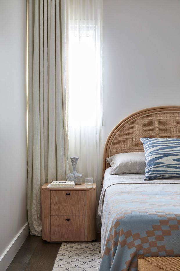 A softly lit bedroom corner featuring a curved rattan headboard, layered white bedding, and a blue-and-white patterned accent pillow and coverlet. A rounded timber bedside table with drawers sits beside the bed, with a small glass lamp and drinking glass on top. Full-height pale curtains frame a bright window, while dark timber flooring and a patterned rug add contrast.