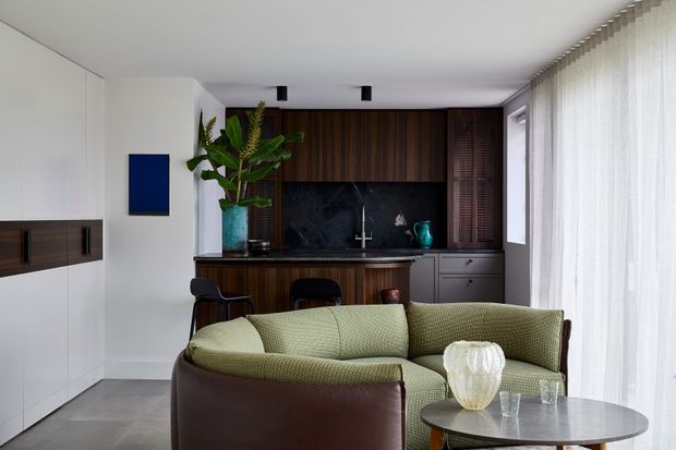 A refined open-plan living area with a compact kitchen behind a curved brown sofa. The room features white built-in cabinetry, grey tiled flooring, a dark timber kitchen wall and island, black stone splashback, grey lower cabinetry, sheer full-height curtains, and contemporary furniture in muted green, brown, and neutral tones.