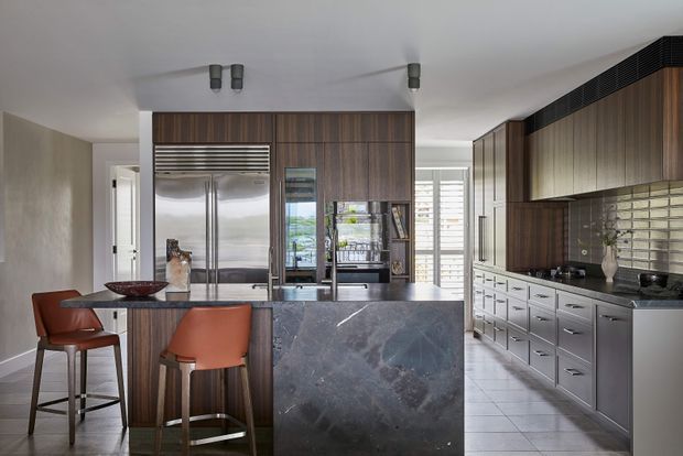 Contemporary kitchen featuring a central island with a dark stone waterfall benchtop, extensive dark timber cabinetry, integrated stainless-steel appliances, and a tiled floor. The space includes a small dining area with two rust-coloured bar stools, reflective backsplash tiles, and ample natural light.
