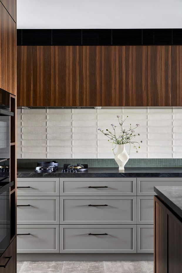 A refined kitchen interior with full-height grey cabinetry, dark stone benchtops, warm vertical timber overhead cabinetry, a black horizontal ventilation grille, and a white textured rectangular-tile splashback. A muted green tile strip runs beneath the splashback, with a gas cooktop and a sculptural white vase on the counter. The room has a balanced, contemporary feel with strong material contrast and integrated joinery.
