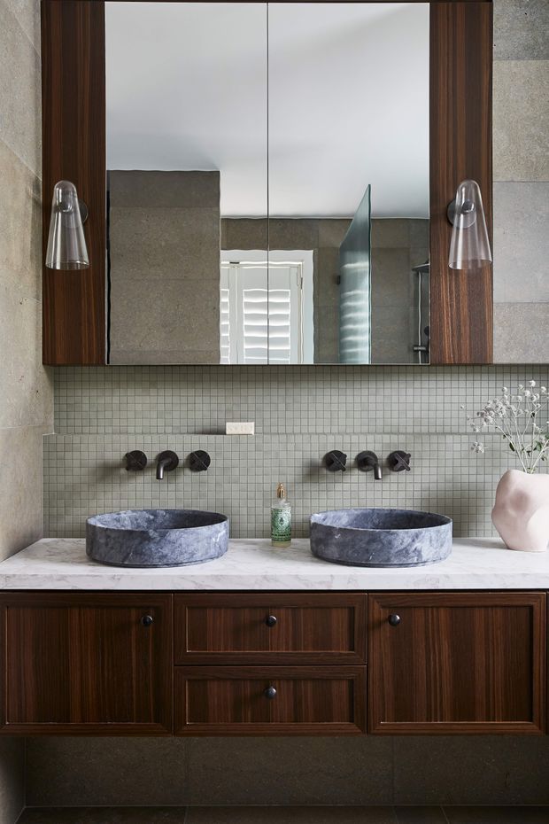 A refined bathroom vanity with two round dark stone vessel basins, wall-mounted black tapware, a pale marble benchtop, dark timber cabinetry, and a small square grey tiled backsplash. A large dark timber-framed mirrored cabinet spans the wall above the vanity, with sculptural wall sconces on either side. The room has a warm, sophisticated, contemporary feel with natural stone and rich wood finishes.