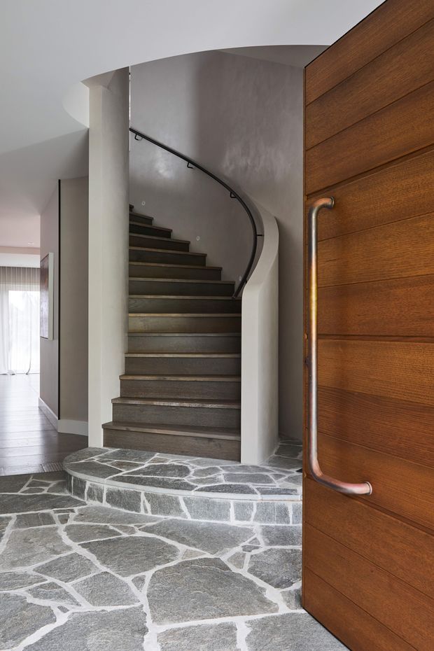 Interior view through an open timber entry door toward a curved stairway. The staircase has dark timber or stone treads, a dark curved metal handrail, and sculpted white plaster walls. The entry floor and raised stair landing are finished in irregular grey stone slabs with light mortar joints. A bright adjoining living area is visible to the left, while the door features horizontal warm wood boards and a tubular metal pull handle.