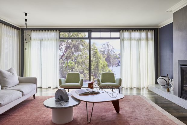 A refined living room with floor-to-ceiling glazing framed by sheer white curtains, a pale upholstered sofa, two green lounge chairs, nested round coffee tables, and a large muted rose rug. A dark blue feature wall with a built-in fireplace contrasts with white marble-look trim and a bright, leafy outlook beyond the windows.