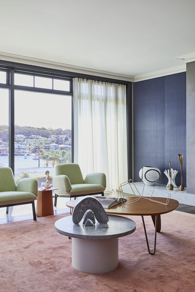 A refined living room with expansive dark-framed corner glazing overlooking a leafy waterfront or suburban view. The room features a deep navy textured feature wall, white ceiling with traditional cornice detailing, sheer curtains, pale green upholstered lounge chairs, sculptural coffee tables, and a muted pink area rug. The overall composition blends contemporary furniture with classic architectural detailing and abundant natural light.