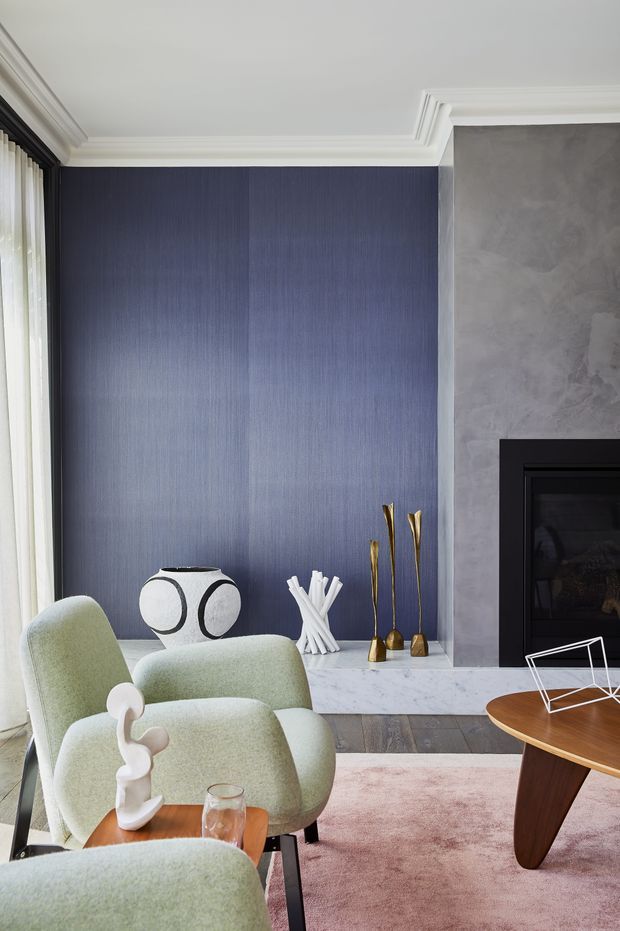 A refined living room featuring a deep blue textured feature wall, pale grey plaster fireplace surround with black firebox, white marble hearth, ornate white ceiling mouldings, and large glazing with sheer curtains. Pale green upholstered armchairs, a sculptural white vase, gold decorative objects, a timber coffee table, and a muted pink rug create a sophisticated contemporary-classic interior.