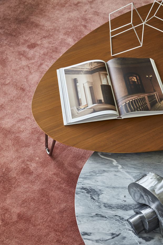 Close-up interior styling vignette showing two overlapping round coffee tables: a warm dark timber table above a veined grey marble table, set on a dusty rose carpet. An open architecture or interior-design book and white geometric wire sculpture sit on the upper table, while a metallic sculptural object rests on the marble table. The image has a refined, editorial, contemporary feel.