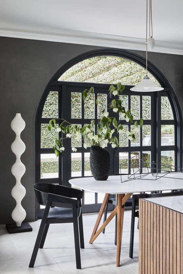 A refined dining area with a large arched, black-framed multi-pane window or glazed door dominating the dark charcoal wall. The space features a white dining table with angled timber legs, black dining chairs, a sculptural white floor lamp, pendant lighting, a dark textured vase with greenery, and a timber-slatted built-in cabinet or island in the foreground. White plaster ceiling cornices and pale flooring create contrast with the dark architectural framing.