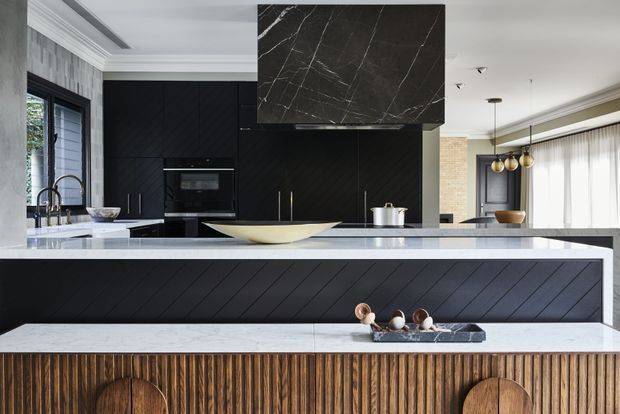 Contemporary open-plan kitchen with a large island and layered breakfast bar, featuring matte black diagonally grooved cabinetry, a dramatic dark veined stone rangehood surround, pale stone benchtops, and a richly textured timber fluted front. A dining area with pendant lighting is visible beyond the kitchen.