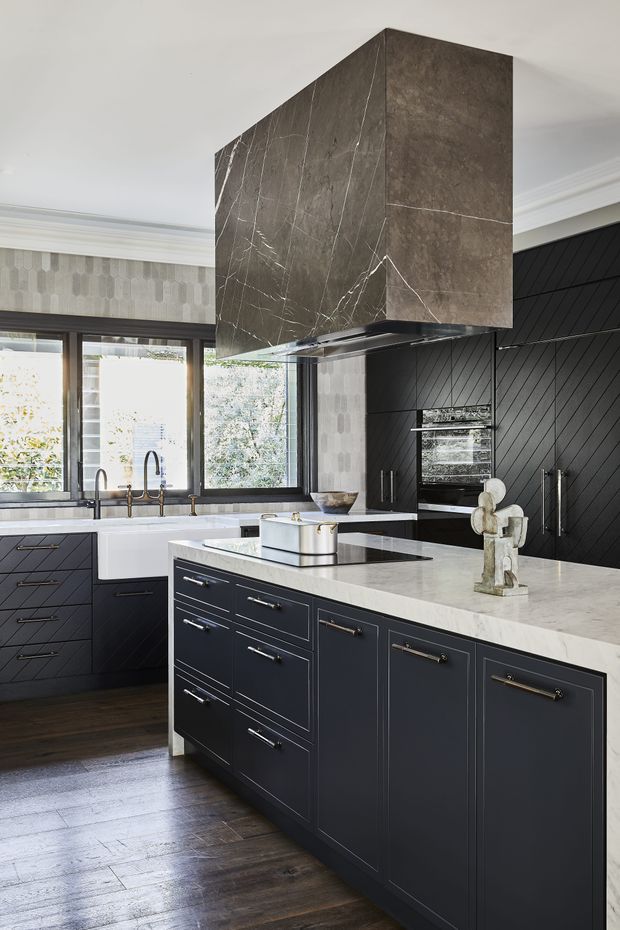A refined, dark-toned kitchen featuring a large central island with pale stone benchtops, deep navy or charcoal cabinetry, brass hardware, a farmhouse-style sink, integrated appliances, and a dramatic marble-look rangehood enclosure. Dark timber flooring, patterned wallcovering, black diagonal wall panelling, and abundant window light create a sophisticated, high-contrast interior.