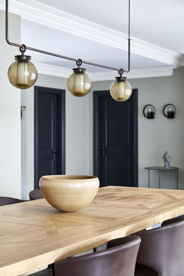 A refined dining room with a large light timber dining table, dark upholstered dining chairs, pale grey walls, black paneled interior doors, ornate white ceiling cornices, and a linear arrangement of three smoked-glass pendant lights with dark metal fittings. A simple ceramic bowl sits on the table, while a slim console and wall sconces appear in the background.