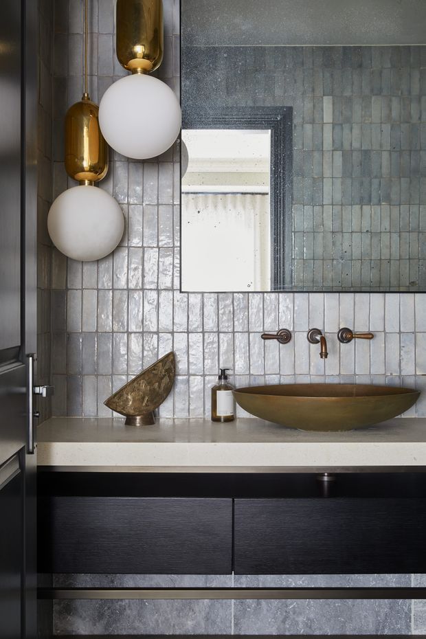 Close-up view of a sophisticated bathroom vanity featuring a wall-mounted black timber-grain cabinet, pale stone benchtop, bronze vessel basin and wall-mounted bronze tapware. The walls are clad in glossy, irregular grey-blue ceramic tiles, complemented by a large dark-framed mirror and sculptural globe pendant lights with brushed brass fittings. The palette is moody and refined with warm metallic accents.
