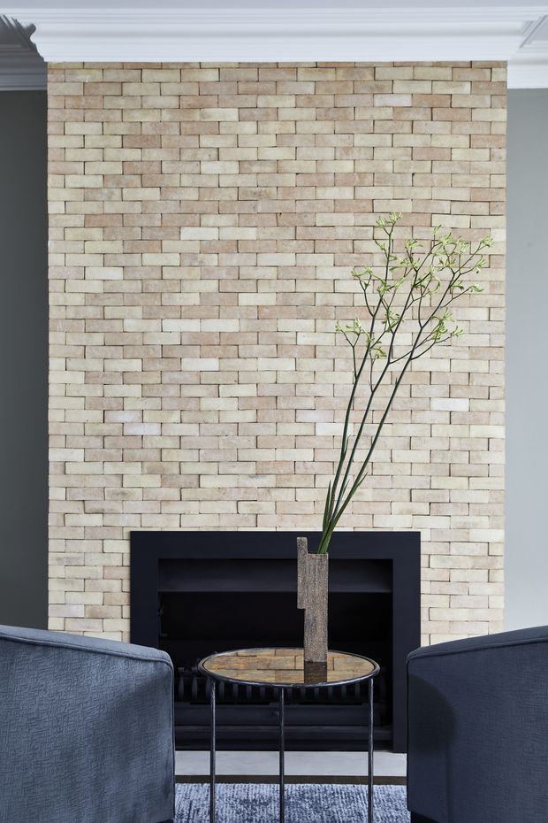 A refined living room vignette centered on a full-height pale brick fireplace wall with crisp white decorative cornice moulding. A dark built-in fireplace surround sits behind a round stone- or tile-topped metal coffee table and tall vase with branches, flanked by two deep blue upholstered armchairs. The room has a restrained, formal palette of cream, white, charcoal, and navy-blue.