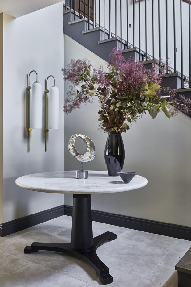A refined interior vignette beneath a staircase, featuring a round white marble-topped pedestal table with a black sculptural base, decorative vase and floral arrangement, metallic circular sculpture, and bowl. The space has pale grey walls, black skirting, grey stone-look flooring, sculptural wall sconces, and a black metal stair balustrade with vertical rods.
