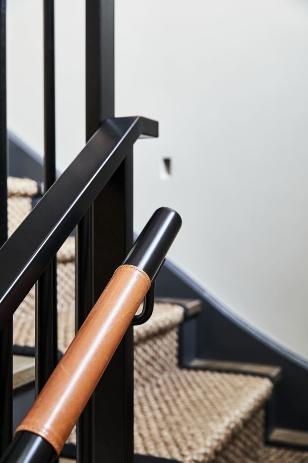 Close-up architectural detail of a contemporary staircase with a black metal balustrade and handrail paired with a warm tan leather-wrapped handrail section. The stair treads are covered in textured beige carpet, with white walls and dark trim creating a crisp, high-contrast interior.