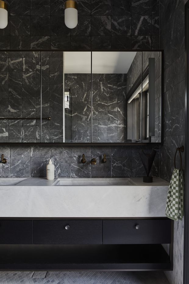 A dark, refined bathroom featuring full-height charcoal marble-look wall tiles with bold white veining, a large mirrored wall cabinet, a pale stone vanity benchtop with integrated basin, dark timber or black cabinetry, and brushed brass wall-mounted tapware and pendant lights. Small-format grey floor tiles and a green-and-white checked hand towel add subtle contrast.