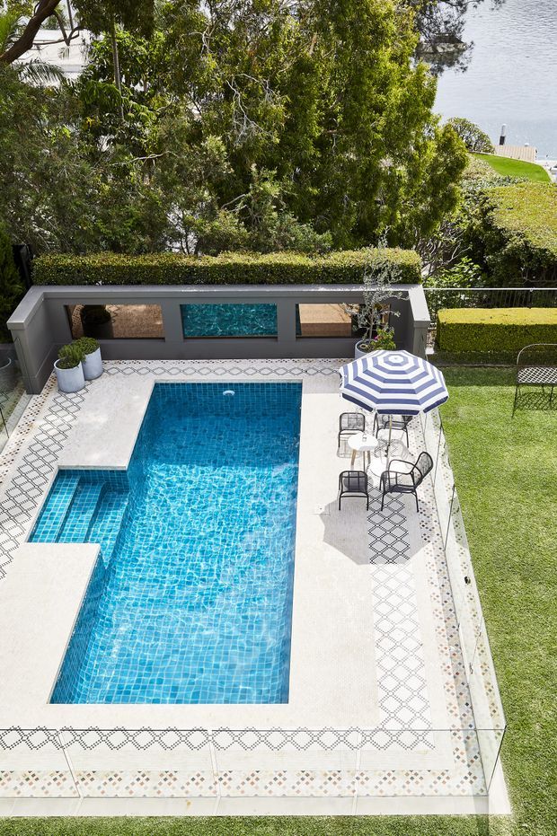 Elevated view of a landscaped residential pool area with a rectangular blue-tiled swimming pool, integrated shallow step section, pale paved surrounds with decorative geometric tile borders, clipped hedges, lawn, outdoor dining furniture and a striped umbrella. A low rendered boundary wall features recessed artwork, with dense greenery and a glimpse of water beyond.