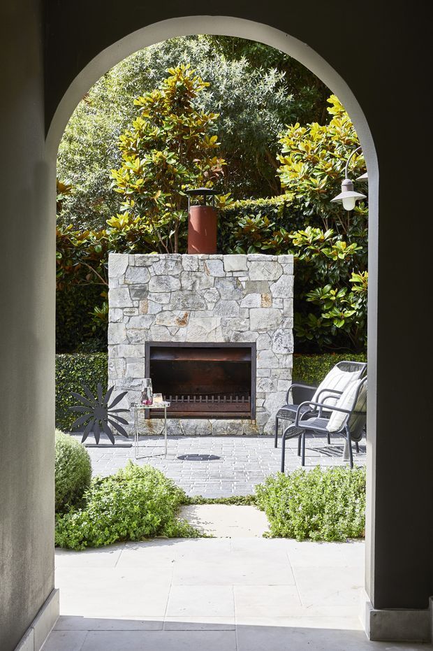A view through a tall rounded archway toward a landscaped outdoor courtyard. The courtyard features a substantial irregular light-stone fireplace with a metal chimney, paved flooring, trimmed hedges and dense planting, plus two outdoor lounge chairs and a small side table. The composition has a Mediterranean-inspired, garden-room feel with strong indoor–outdoor connection.