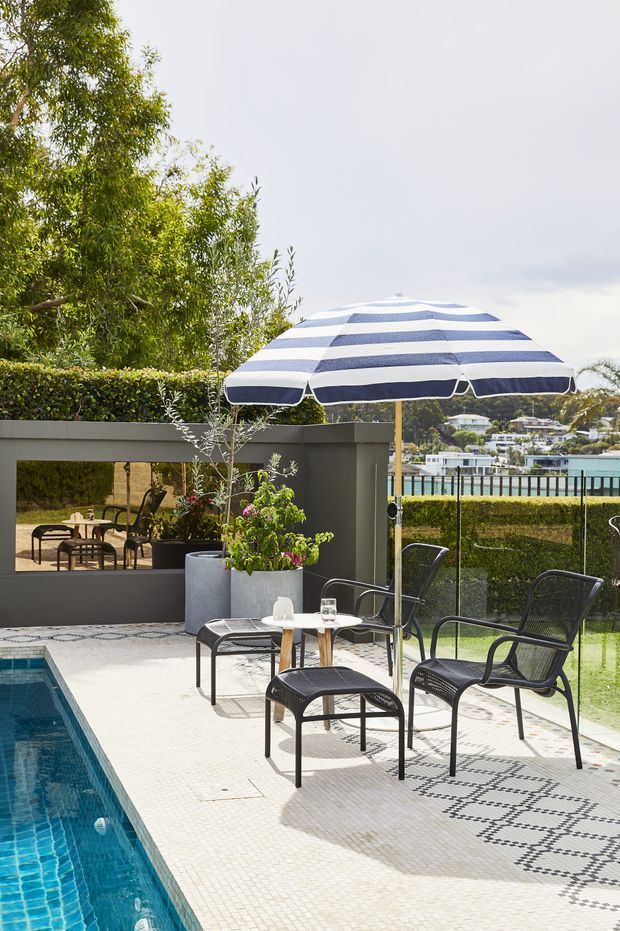 A bright outdoor poolside terrace with a blue-and-white striped patio umbrella, black metal outdoor lounge chairs and table, large planters, patterned pale tile paving, and a swimming pool at left. A low wall and garden screening frame views toward a waterfront residential setting in the background.